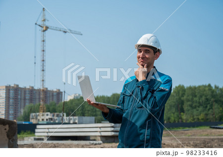 A male builder in a helmet stands against the background of a construction site with a laptop in his hands. A male builder in a helmet stands against the background of a construction site with a laptop in his hands. 98233416