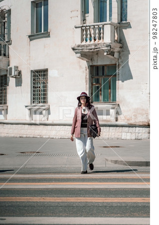 Woman city road crossing. Stylish woman in a hat crosses the road at a pedestrian crossing in the city. Dressed in white trousers and a jacket with a bag in her hands. Woman city road crossing. Stylish woman in a hat crosses the road at a pedestrian crossing in the city. Dressed in white trousers and a jacket with a bag in her hands. 98247803