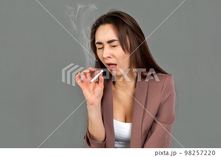 A young woman in a jacket crouches and tries to smoke a cigarette. Gray background. Copy space. The concept of nicotine addiction and Smoking control 98257220