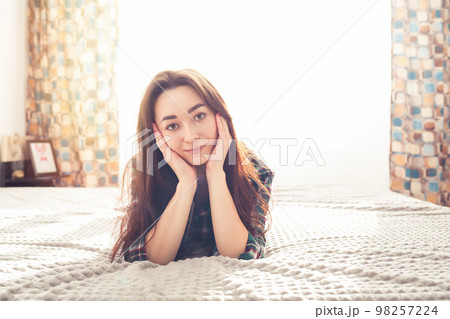 Portrait of a young beautiful brunette lying on the bed, and leaning her face on her chin. In the background, a bright room, a window and light. Copy space Portrait of a young beautiful brunette lying on the bed, and leaning her face on her chin. In the background, a bright room, a window and light. Copy space 98257224