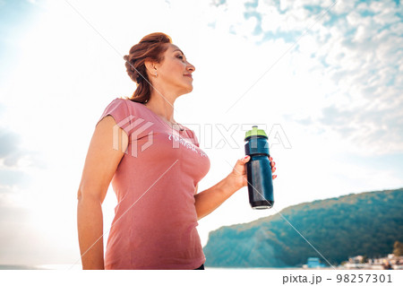 An adult woman in sportswear, smiling and holding a shaker in her hands. In the background, the sky and mountains. Bottom view. The concept of sports lifestyle An adult woman in sportswear, smiling and holding a shaker in her hands. In the background, the sky and mountains. Bottom view. The concept of sports lifestyle 98257301