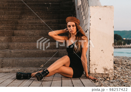 Sexy blonde in a black dress and hat posing sitting on the steps. In the distance you can see the sea. Close up 98257422