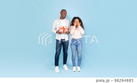 Greeting concept. Loving black man holding gift box, excited lady covering her eyes with hands and waiting for surprise 98264885
