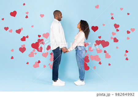 Full length shot of happy black couple holding hands and looking at each other on blue background with red flying hearts 98264992