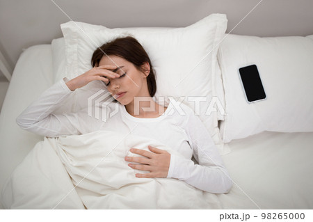 Upset young woman lying in bed with smartphone on pillow 98265000