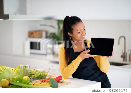 Happy young african american woman in apron show tablet with empty screen at table with fresh vegetables 98265175