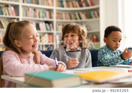 Group of funny diverse international pupils talking and laughing, sitting at table with notebooks and books in classroom 98265265