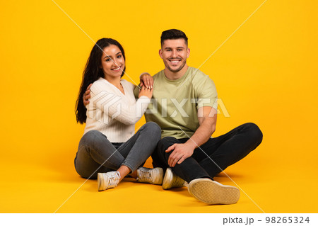 Cheerful millennial arab guy and lady in casual sit on floor, enjoy date, isolated on orange background, studio Cheerful millennial arab guy and lady in casual sit on floor, enjoy date, isolated on orange background, studio 98265324