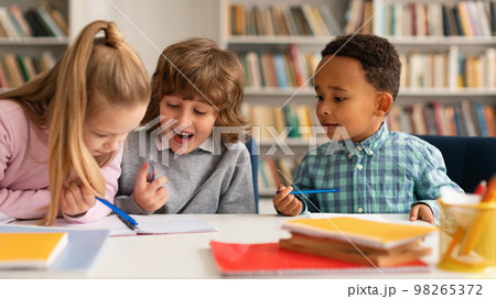 School time. Multiethnic kids writing in notebooks and smiling, talking with each other and laughing, sitting at table 98265372