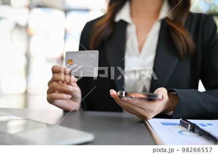 Businesswoman sits at her desk holding her phone and showing her credit card to camera 98266557