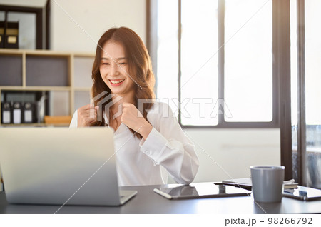 Cheerful young Asian female office worker looking at laptop screen, celebrating her success Cheerful young Asian female office worker looking at laptop screen, celebrating her success 98266792