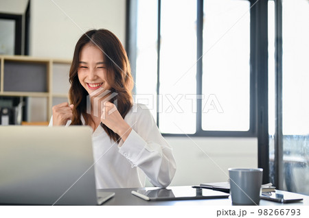 Successful and cheerful young Asian female office worker looking at laptop screen, celebrating Successful and cheerful young Asian female office worker looking at laptop screen, celebrating 98266793