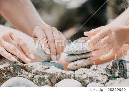 Woman with daughter bilds stones pyramid on seashore on a sunny day on the blue sea background. Happy family holidays. Pebble beach, calm sea. Concept of happy vacation on the sea, meditation, spa 98269040