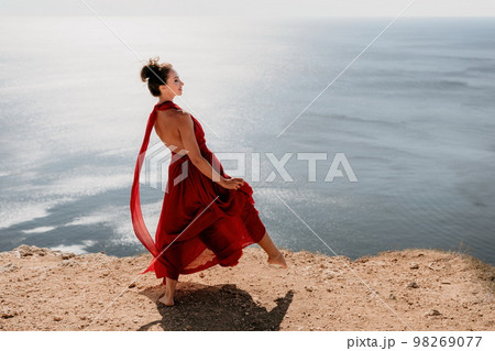 Woman in red dress on sea. Side view a Young beautiful sensual woman in a red long dress posing on a rock high above the sea on sunset. Girl on the nature on blue sky background. Fashion photo. Woman in red dress on sea. Side view a Young beautiful sensual woman in a red long dress posing on a rock high above the sea on sunset. Girl on the nature on blue sky background. Fashion photo. 98269077