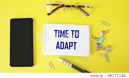Time to adapt text written on notepad and yellow background with mobile phone and pen 98272186