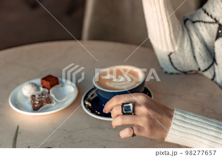 A cup of fresh cappuccino coffee in the hands of a woman on a fashionable background of a white marble table, next to a plate with sweets. Coffee addiction A cup of fresh cappuccino coffee in the hands of a woman on a fashionable background of a white marble table, next to a plate with sweets. Coffee addiction 98277657