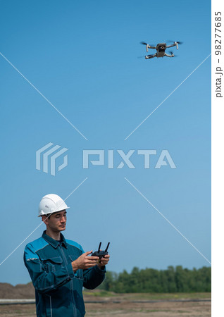 A man in a helmet and overalls controls a drone at a construction site. The builder carries out technical oversight. 98277685