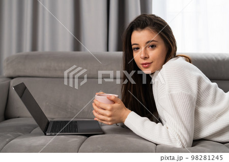 Portrait of a beautiful young girl lying on a sofa with a cup of tea and a laptop. Portrait of a beautiful young girl lying on a sofa with a cup of tea and a laptop. 98281245
