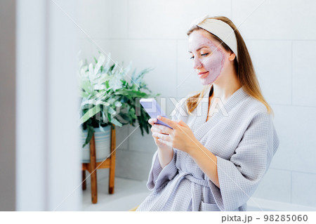 Smiling woman with facial pink clay mask on the face using smartphone at home bath enjoying relaxation and spa beauty treatment. Teen Beauty blogger creating content. Selective focus, copy space Smiling woman with facial pink clay mask on the face using smartphone at home bath enjoying relaxation and spa beauty treatment. Teen Beauty blogger creating content. Selective focus, copy space 98285060