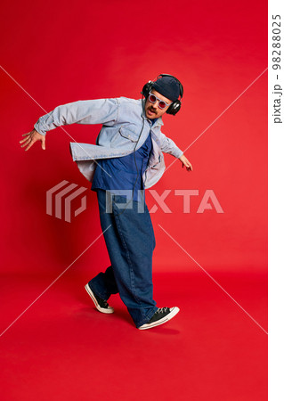 Hip hop. Portrait of man in stylish clothes, oversized jeans, shirt and hat posing, dancing over red background. Concept of modern fashion, lifestyle, music culture 98288025