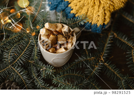 A mug of hot chocolate with cinnamon and marshmallows surrounded by fir branches, garlands and a yellow-blue knitted hat. 98291525