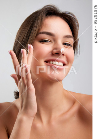 Close up beauty portrait of happy beautiful woman with bared shoulders applying face cosmetics products and looking with delighted facial expression isolated over light background 98291876