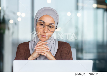 Serious bored businesswoman inside office, muslim woman in hijab thinking while sitting at workplace with laptop, woman at work thinking about decisions. Serious bored businesswoman inside office, muslim woman in hijab thinking while sitting at workplace with laptop, woman at work thinking about decisions. 98294632