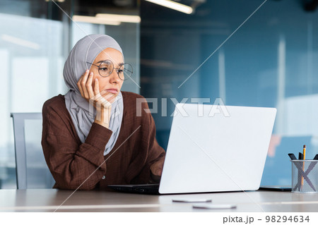 Serious bored businesswoman inside office, muslim woman in hijab thinking while sitting at workplace with laptop, woman at work thinking about decisions. Serious bored businesswoman inside office, muslim woman in hijab thinking while sitting at workplace with laptop, woman at work thinking about decisions. 98294634