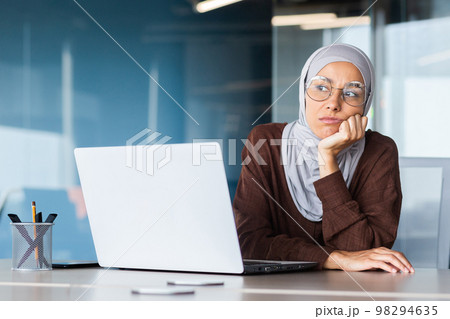Serious bored businesswoman inside office, muslim woman in hijab thinking while sitting at workplace with laptop, woman at work thinking about decisions. 98294635