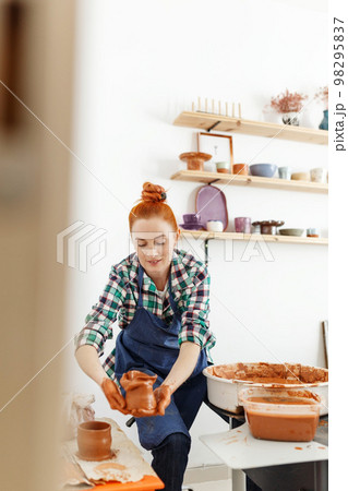Cheerful female sculptor while she making clay pot on pottery wheel. Idea of small business and entrepreneurship. Home hobby, entertainment and leisure. Art studio Cheerful female sculptor while she making clay pot on pottery wheel. Idea of small business and entrepreneurship. Home hobby, entertainment and leisure. Art studio 98295837