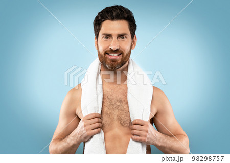 Portrait of happy shirtless bearded man with towel on neck posing on blue studio background and smiling at camera 98298757