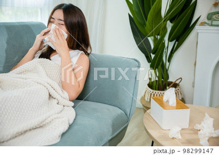 Asian young woman she sick blowing nose sneezing in tissue at home, fever caught cold, Sick female sitting under blanket on sofa and sneeze with tissue paper in living room, medicine healthcare Asian young woman she sick blowing nose sneezing in tissue at home, fever caught cold, Sick female sitting under blanket on sofa and sneeze with tissue paper in living room, medicine healthcare 98299147