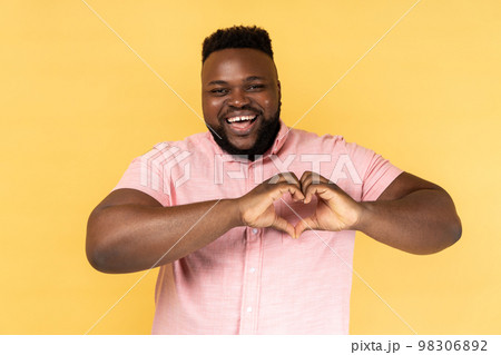 Portrait of lovely romantic happy man wearing pink shirt making heart shape with fingers, gesturing love hope charity sign, looks at camera. Indoor studio shot isolated on yellow background. 98306892