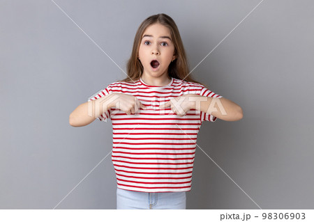 Portrait of astonished little girl wearing striped T-shirt pointing at herself, asks who me, has surprised expression, shocked being picked. Indoor studio shot isolated on gray background. 98306903