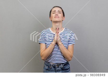 Please, I'm begging. Portrait of upset worried woman wearing striped T-shirt looking up with imploring desperate grimace, praying to god asking for help. Indoor studio shot isolated on gray background Please, I'm begging. Portrait of upset worried woman wearing striped T-shirt looking up with imploring desperate grimace, praying to god asking for help. Indoor studio shot isolated on gray background 98306949