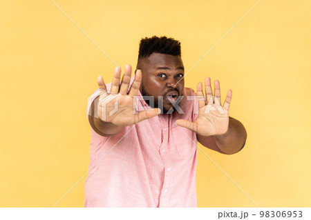 No, it's scary. Portrait of frightened shocked man wearing pink shirt raising hands in fear, looking horrified and panicking, hiding from phobia. Indoor studio shot isolated on yellow background. 98306953