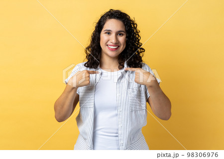 Portrait of friendly woman with dark wavy hair pointing finger at herself feeling proud, ambitious, boasting successful achievement. Indoor studio shot isolated on yellow background. 98306976