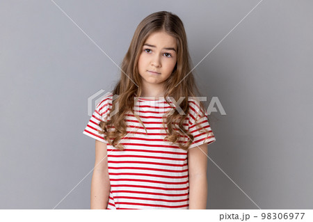 Portrait of unhappy little girl wearing striped T-shirt being upset of bad news, looking at camera with frowning face, expressing sadness. Indoor studio shot isolated on gray background. 98306977