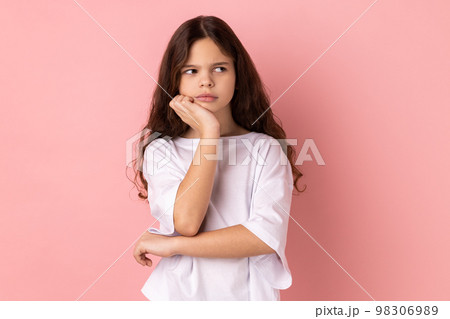 Portrait of thoughtful pensive little girl wearing white T-shirt thinking about future, holding chin, having serious facial expression. Indoor studio shot isolated on pink background. 98306989