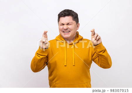 Portrait of middle aged man crossing fingers and closing eyes making wish, believing in miracle, hope in success, wearing urban style hoodie. Indoor studio shot isolated on white background. 98307015