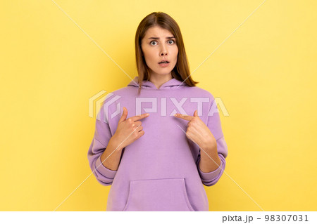 Portrait of amazed woman pointing herself and looking at camera with shocked expression, astonished by sudden success, wearing purple hoodie. Indoor studio shot isolated on yellow background. 98307031