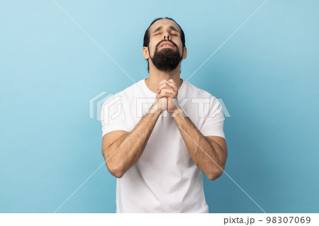 Man wearing white T-shirt looking up holding hands together, praying God, asking for health and support pleading for healing and forgiveness. Indoor studio shot isolated on blue background. 98307069