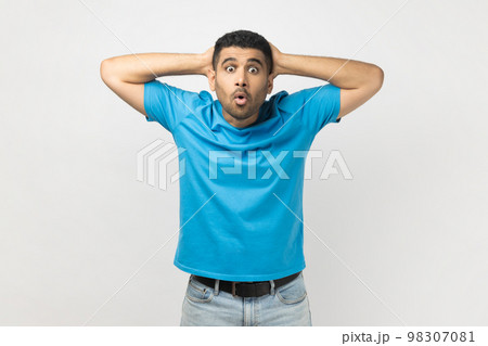 Portrait of shocked surprised man wearing blue T- shirt standing looking at camera with big eyes, keeps hands behind head, sees something astonished. Indoor studio shot isolated on gray background. Portrait of shocked surprised man wearing blue T- shirt standing looking at camera with big eyes, keeps hands behind head, sees something astonished. Indoor studio shot isolated on gray background. 98307081
