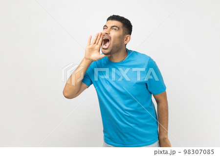 Portrait of excited handsome attractive unshaven man wearing blue T- shirt standing keeps hands near opened mouth, screaming news. Indoor studio shot isolated on gray background. 98307085