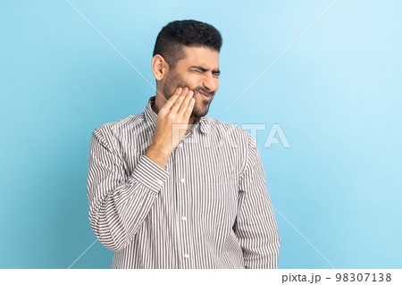 Unhappy bearded businessman feeling toothache, touching sore cheek, suffering from cavities, cracked teeth, gum recession, wearing striped shirt. Indoor studio shot isolated on blue background. Unhappy bearded businessman feeling toothache, touching sore cheek, suffering from cavities, cracked teeth, gum recession, wearing striped shirt. Indoor studio shot isolated on blue background. 98307138