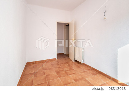 An empty spacious room with an open door, white walls and ceiling, caramel-colored square floor tiles and a radiator on the wall for warmth in the cold season. The room is lit by bright daylight. 98312013