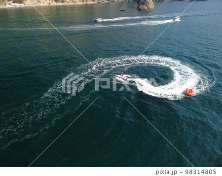 Happy people swim on air mattress behind a high speed water bike. Tourists ride on inflatable watercraft boat. Sea attraction. Water scooter skating people on large Inflatable raft. Aerial copter view 98314805