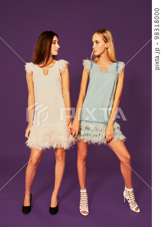 Professional photo of two young women, blonde and brunette, in cocktail dresses posing on purlpe background Professional photo of two young women, blonde and brunette, in cocktail dresses posing on purlpe background 98318000