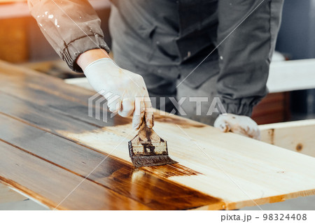 Man paints wooden boards with paint brush. Carpenter cabinetmaker varnishes wooden surface 98324408