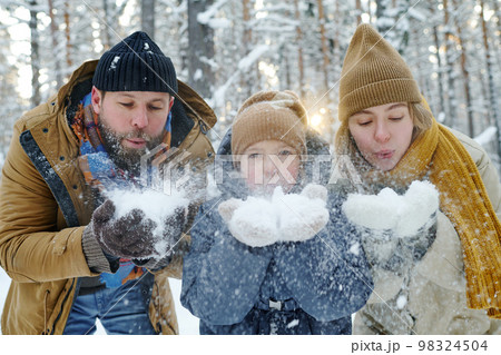 Portrait of happy family of three blowing snow from their hands during walk in the park 98324504